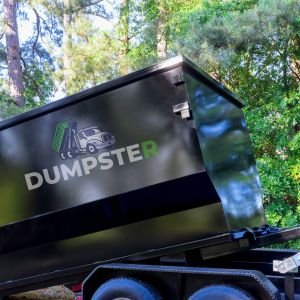Black Roofing Dumpster A black dumpster alongside a black trailer, set against a backdrop of trees in a quiet woodland.